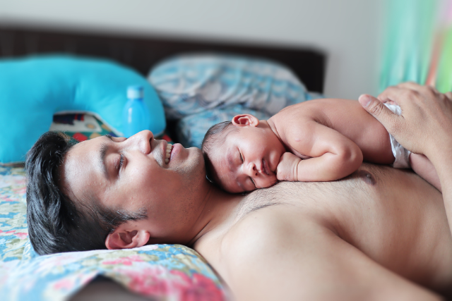 An East Asian man lays on bed with a newborn baby on his bare chest on the Informed Pregnancy Blog