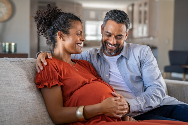 A pregnant woman and a man sit on the couch smiling, they each have a hand on the woman's round belly on The informed Pregnancy Blog.